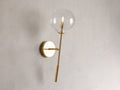 Front view of the brass Roscoe Sconce, featuring a clear glass globe with an illuminated LED bulb, mounted on a wall.