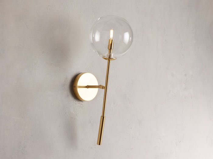 Front view of the brass Roscoe Sconce, featuring a clear glass globe with an illuminated LED bulb, mounted on a wall.