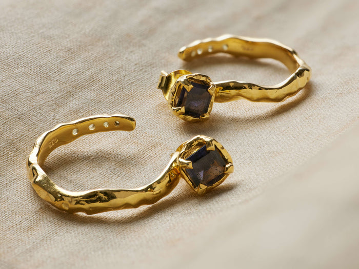 Two Safiya Earrings, gold-plated with hammered texture, feature dark square faceted peridot stones, on light textured fabric.