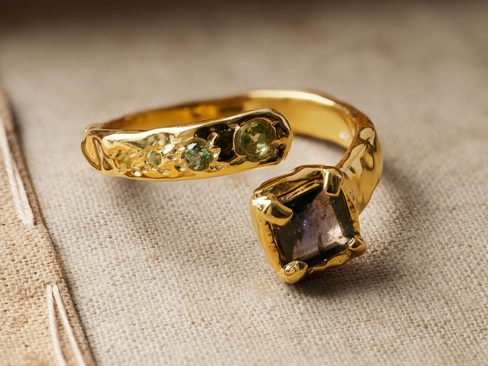 Safiya Ring: Gold-plated, open-ended ring with a textured band, small green peridots, and a large square purple-grey stone.