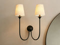 Saige Sconce with two off-white shades & dark metal arms, illuminated on a warm-toned wall next to a mirror.