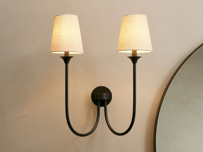 Saige Sconce with two off-white shades & dark metal arms, illuminated on a warm-toned wall next to a mirror.