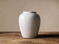 White Santorini Urn with crackle glaze, standing upright on a distressed wooden table against a light tan wall.