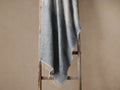 Saraya Throw, a soft, fuzzy light blue/grey blanket with fringed edges, draped over a rustic wooden ladder against a neutral wall.