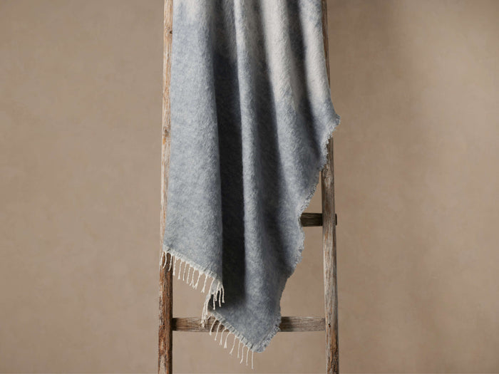 Saraya Throw, a soft, fuzzy light blue/grey blanket with fringed edges, draped over a rustic wooden ladder against a neutral wall.