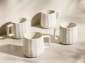 Front view of four cream Saskia Mugs, each featuring a distinct vertical ribbed texture.