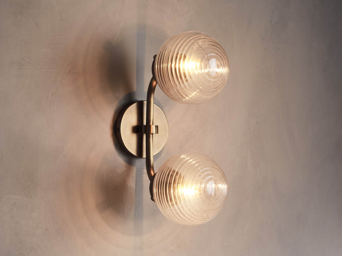 Sayre Double Sconce in plated Burnished Brass with two ribbed glass globes, mounted horizontally, emitting warm light.
