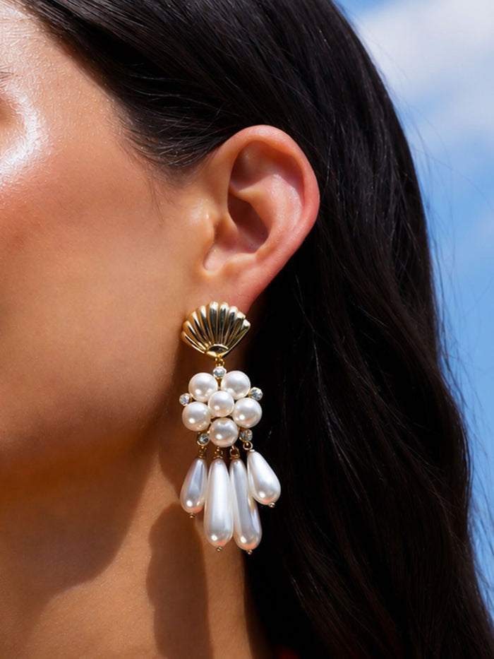 A close-up of a model's ear wearing a dangling earring with a gold scallop-shaped top, a cluster of white pearls, and three teardrop pearl drops against a blue sky background.
