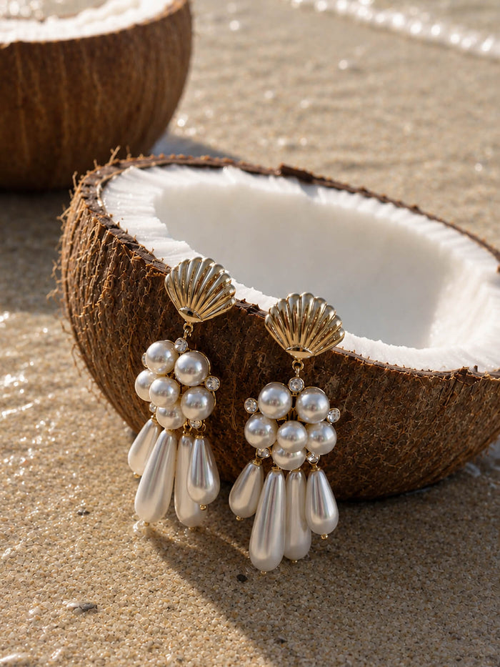 A pair of gold and pearl drop earrings displayed on a halved coconut shell placed on sandy ground, with a blurred coconut and string beads in the background.
