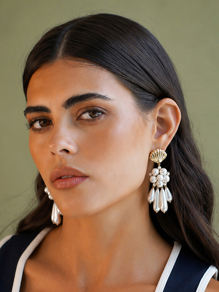 A close-up of a woman wearing large shell-shaped dangle earrings with white pearl-like clusters and silver-toned teardrop drops against a muted olive background.