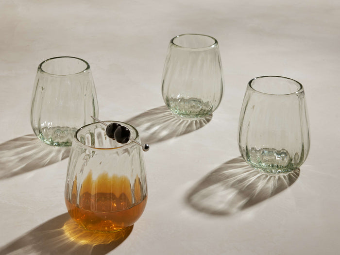 Four clear, textured Seema Double Old-Fashioned Glasses on a light surface. One holds amber liquid with two dark cherries.