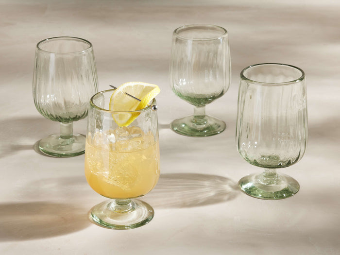 Four ribbed Seema Clear Goblet Glasses with a green tint. One glass in front holds a yellow drink with ice & lemon slice.
