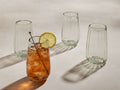 Seema clear highball glasses, set of 4. One glass contains iced amber drink with lemon & stirrer. Textured, recycled glass.