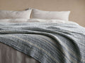 Selva Bed Blanket, blue-grey and white striped, textured boucl?? blanket draped casually over a bed with pillows.