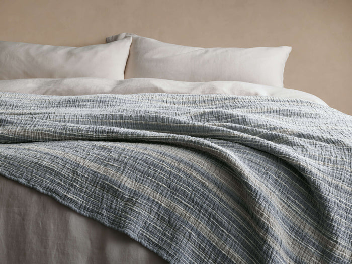 Selva Bed Blanket, blue-grey and white striped, textured boucl?? blanket draped casually over a bed with pillows.