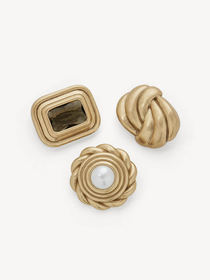 Three gold-tone jewelry pieces arranged on a light gray background: a rectangular bezel-set piece with a dark stone, a twisted knot-shaped piece, and a rope-textured piece with a white pearl center.
