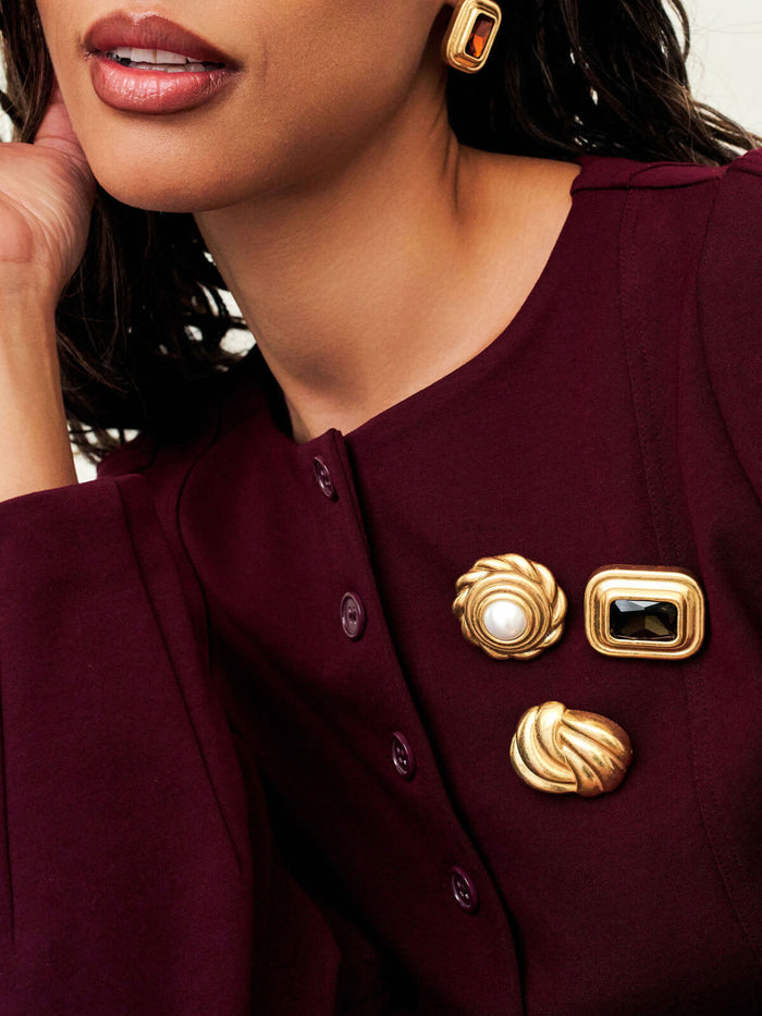 A close-up of a model wearing a deep burgundy top with three decorative gold-tone buttons, including one with a white pearl center, and matching gold earrings.