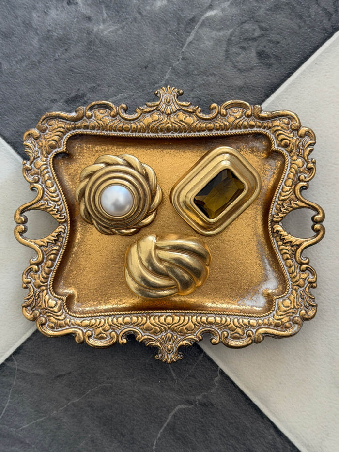 A vintage ornate gold brooch featuring three inset elements: a round pearl-like center, a rectangular amber stone, and two twisted gold floral shapes, displayed on a dark gray and white marble surface.