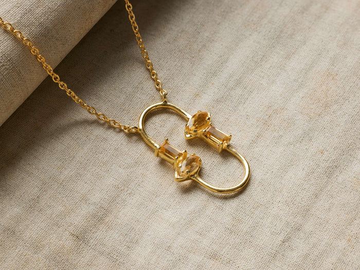 Sienna Citrine Necklace: Gold chain, central pendant with two interconnected loops, each holding two golden citrine stones, on light fabric.