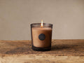 Signature Candle in Sandalwood Leaf and Tobacco, lit. Light brown wax in walnut-tinted glass with black logo on a rustic wooden surface.