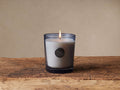 Lit Signature Suede Wood & Vetiver candle in a gray-tinted glass jar with a metallic emblem, on rustic wood.