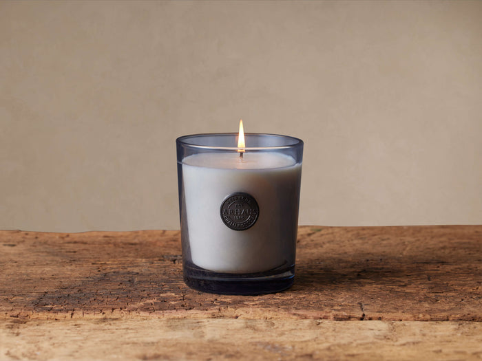 Lit Signature Suede Wood & Vetiver candle in a gray-tinted glass jar with a metallic emblem, on rustic wood.