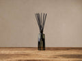 Signature Diffuser: Ribbed glass bottle with subtle yellow tint holds black reeds, standing on a rustic wooden table.