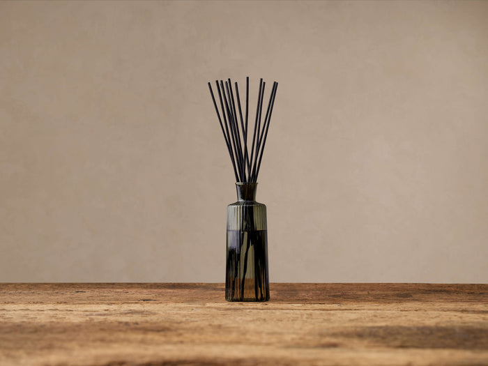 Signature Diffuser: Ribbed glass bottle with subtle yellow tint holds black reeds, standing on a rustic wooden table.