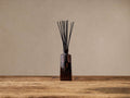 Signature Diffuser: Upright, ribbed walnut brown glass bottle with black reeds on a rustic wooden table against a light beige wall.