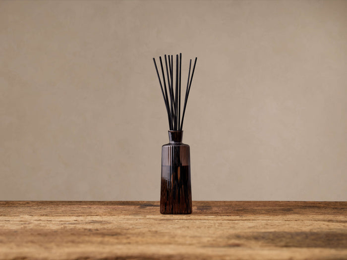 Signature Diffuser: Upright, ribbed walnut brown glass bottle with black reeds on a rustic wooden table against a light beige wall.