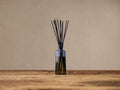 Signature Diffuser: Ribbed gray glass bottle with fanned black reeds, on a rustic wood surface against a beige wall.