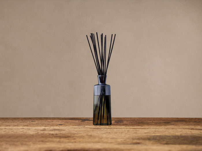 Signature Diffuser: Ribbed gray glass bottle with fanned black reeds, on a rustic wood surface against a beige wall.