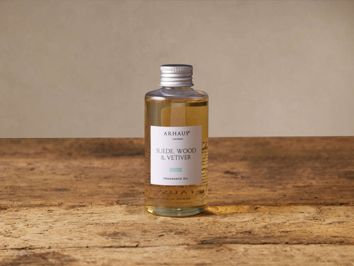Arhaus Signature Suede Wood & Vetiver Diffuser Refill. Clear bottle with amber liquid, silver cap, on rustic wood, light background.