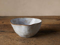 Skye Cereal Bowl: Textured light gray stoneware, irregular shape, dark brown rim. Sits on a rustic wooden table.