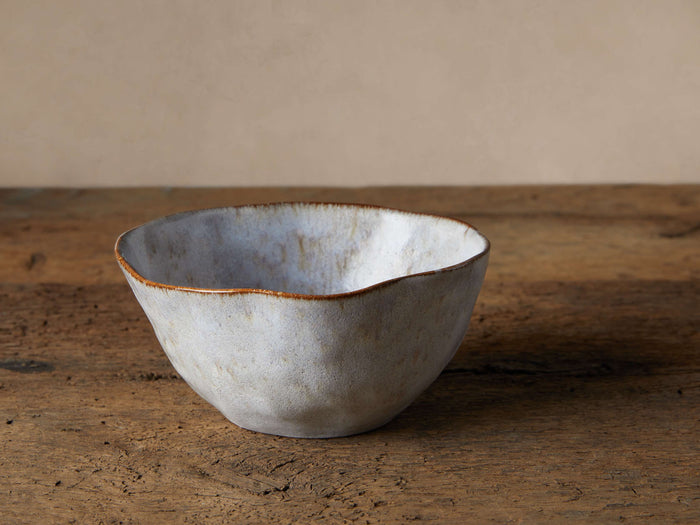 Skye Cereal Bowl: Textured light gray stoneware, irregular shape, dark brown rim. Sits on a rustic wooden table.
