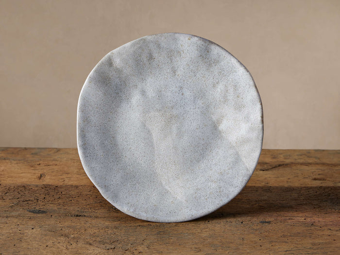 Skye Dinner Plate, light grey reactive glazed stoneware with speckled texture, irregular round shape, standing on a rustic wood surface.