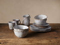 Skye Dinnerware set of 16, white glazed stoneware mugs, bowls, and plates with dark trim, on a rustic wooden table.