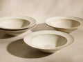 Four off-white deep ceramic dinnerware bowls with wide flat rims and tan speckles, two stacked.