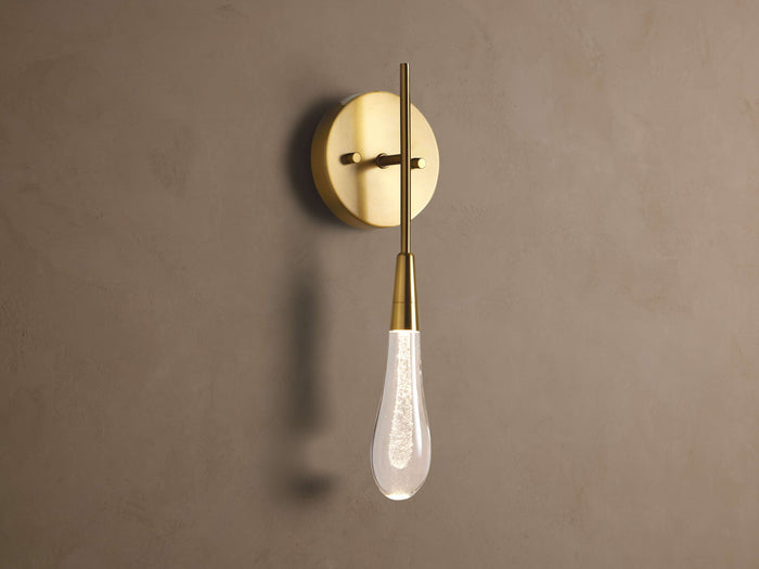 Soltaire Sconce: Round brass backplate, vertical arm, and glowing teardrop glass shade with internal bubbles mounted on a wall.