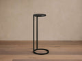 Soma Martini Table with a sleek black metal frame, round top, and circular base, standing on a wooden floor against a beige wall.