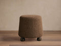 Steffi Ottoman, a brown, tapered bouclé-textured ottoman with three dark chestnut wood legs on a wooden floor.