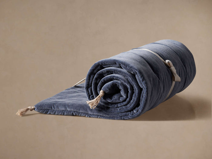Muted blue Stone Washed Velvet Throwbed, partially rolled, with a cream strap and jute tassels, on a beige background.