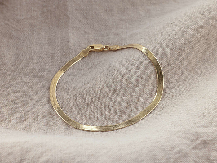 Sura Snake Chain Bracelet in gold vermeil, lying on beige fabric. Features a flat, gleaming design with a secure clasp.