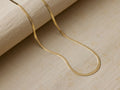 18k gold vermeil Sura Snake Chain Necklace, flat and sleek, gracefully curved on a textured light beige fabric.