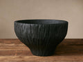 Hand-carved Surma Vessel of black wood with rough, textured surface and tapering shape, sitting on a wooden table.