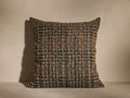 A square cushion with a textured, woven pattern in earthy tones of brown and black, placed on a neutral surface and background.