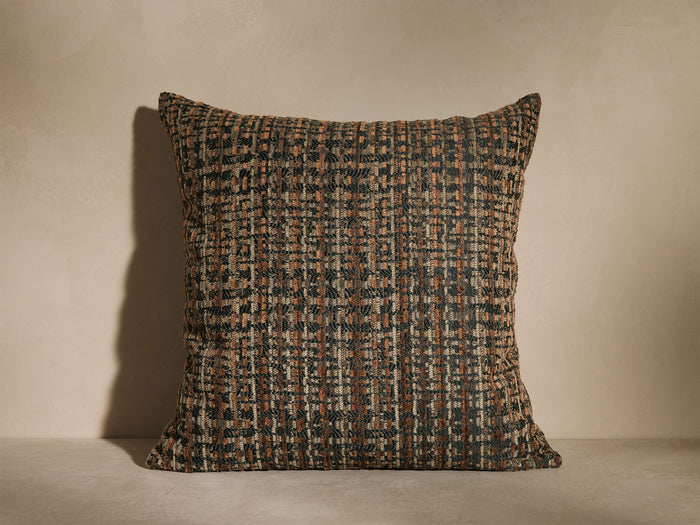 A square cushion with a textured, woven pattern in earthy tones of brown and black, placed on a neutral surface and background.