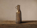 Taddeo Tall Jug, cast aluminum with an antique brass finish, featuring a C-handle, on a rustic wood surface.
