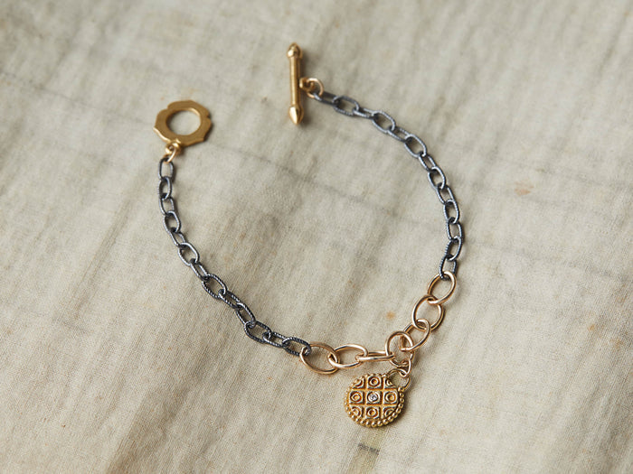 Yellow bronze Tavi bracelet with white diamond charm. Oxidized silver chain, gold-fill links, toggle clasp, on a fabric surface.