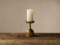 Thira Pillar Holder: an antique-inspired, textured brass candle holder with a lit cream pillar candle, on a rustic wooden table.